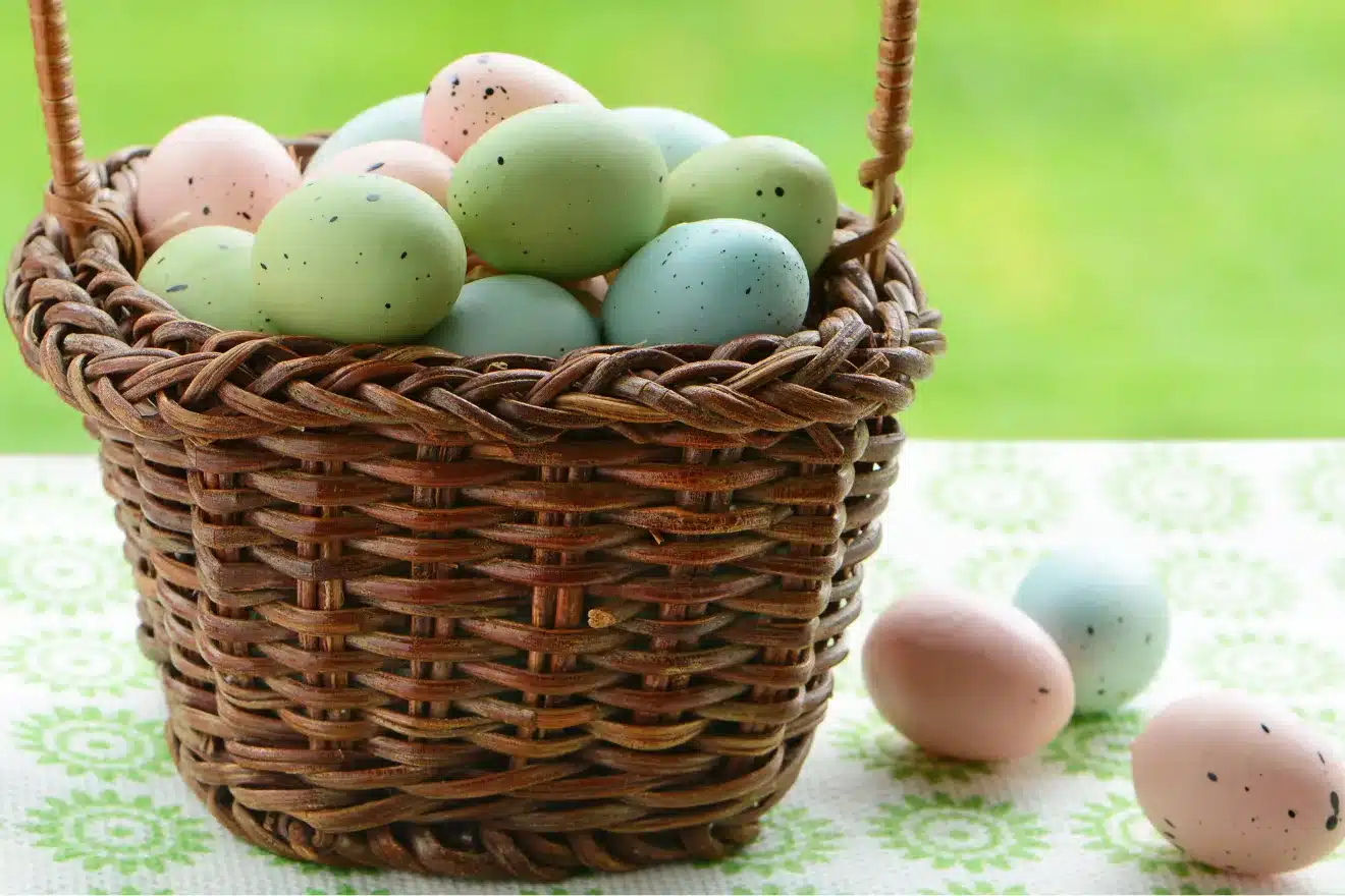 Wicker basket filled with pastel-colored eggs on a patterned tablecloth, with a green background.