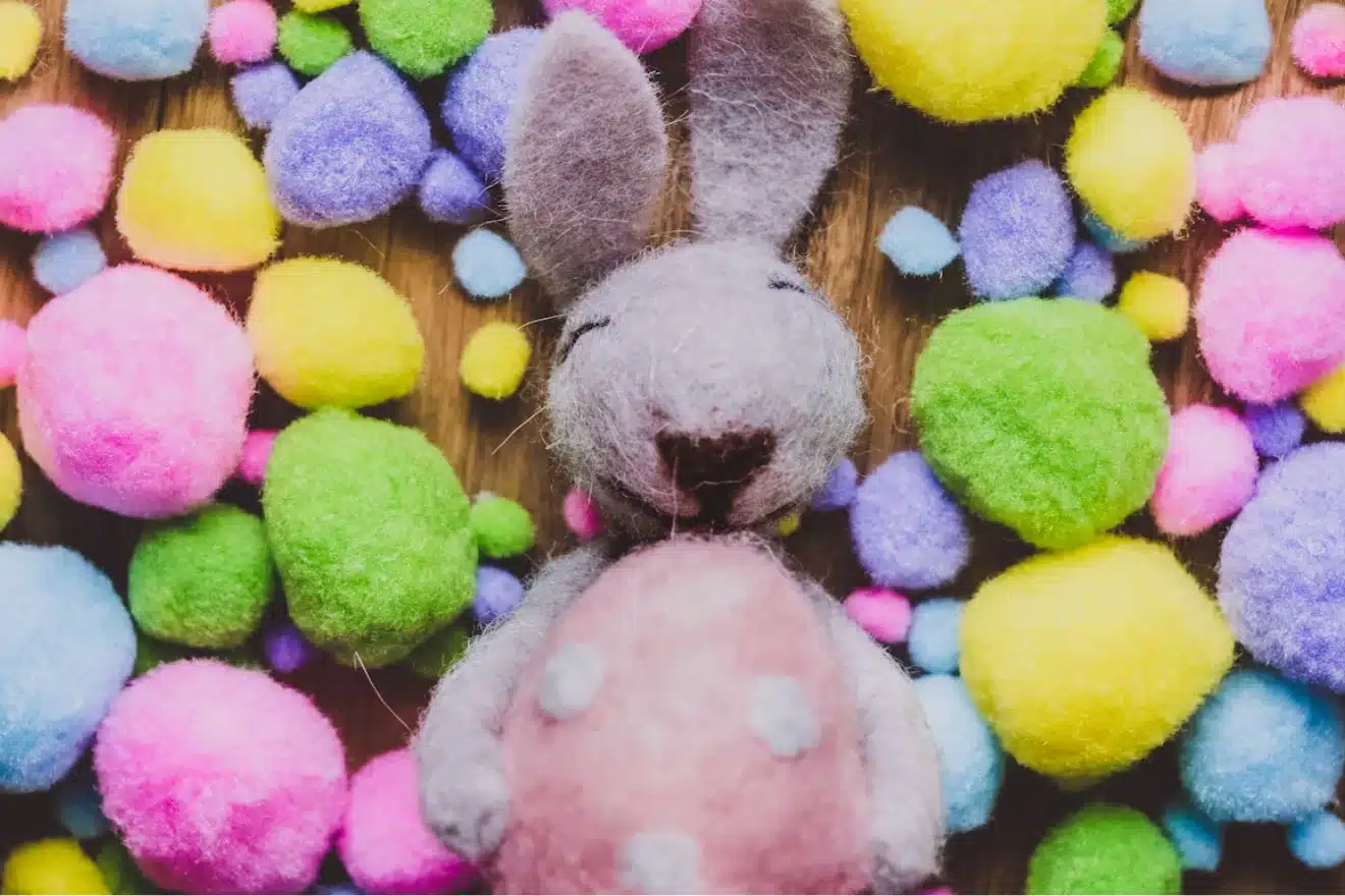 A stuffed gray rabbit with closed eyes and a pink polka-dotted belly is surrounded by colorful pom-poms.