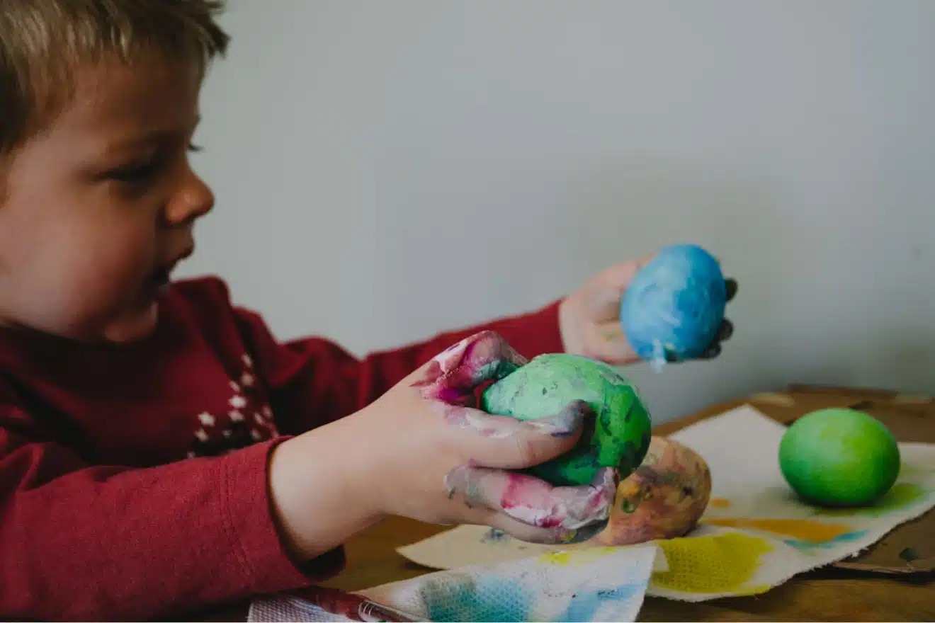 Child with paint-covered hands holding and painting colorful eggs at a table.