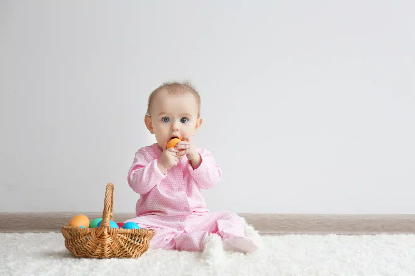 A baby in pink pajamas sits on a white carpet, holding a colored egg with a basket of more eggs nearby.
