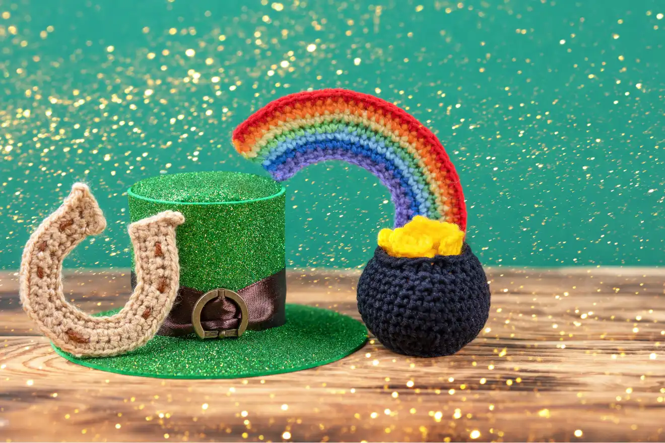 Green glittery hat, crocheted horseshoe, and rainbow with a pot of gold sit on a wooden surface against a green background with golden sparkles.
