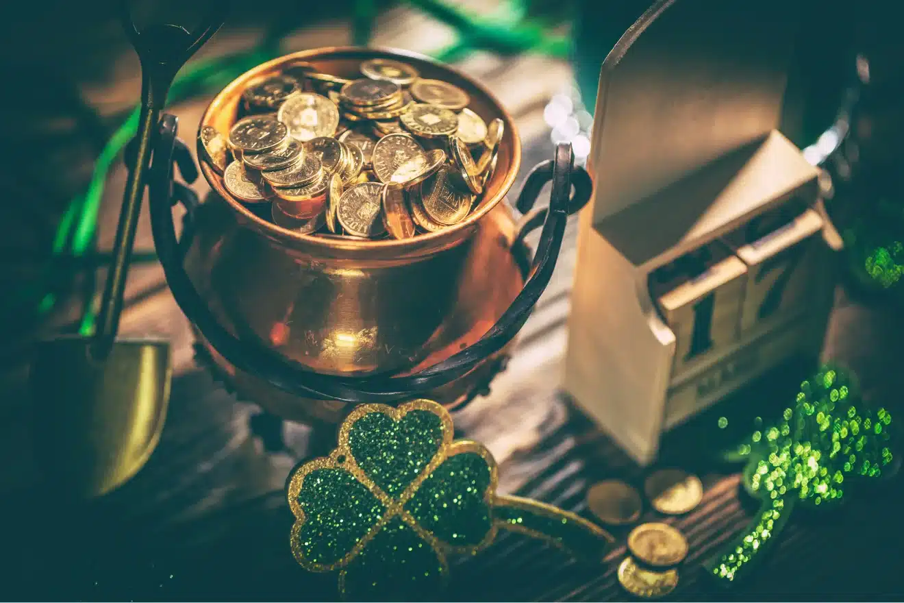 A pot of gold coins, a small shovel, a wooden calendar displaying 17, and a glittery green shamrock are arranged on a wooden surface.