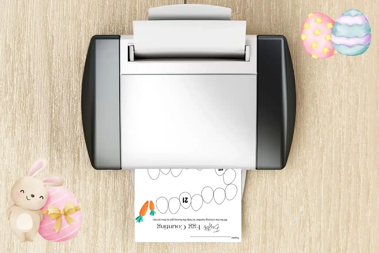 A printer on a wooden surface prints a page featuring a numbered egg pattern with four carrots. Nearby are two decorative eggs and a stuffed toy rabbit holding a striped egg.