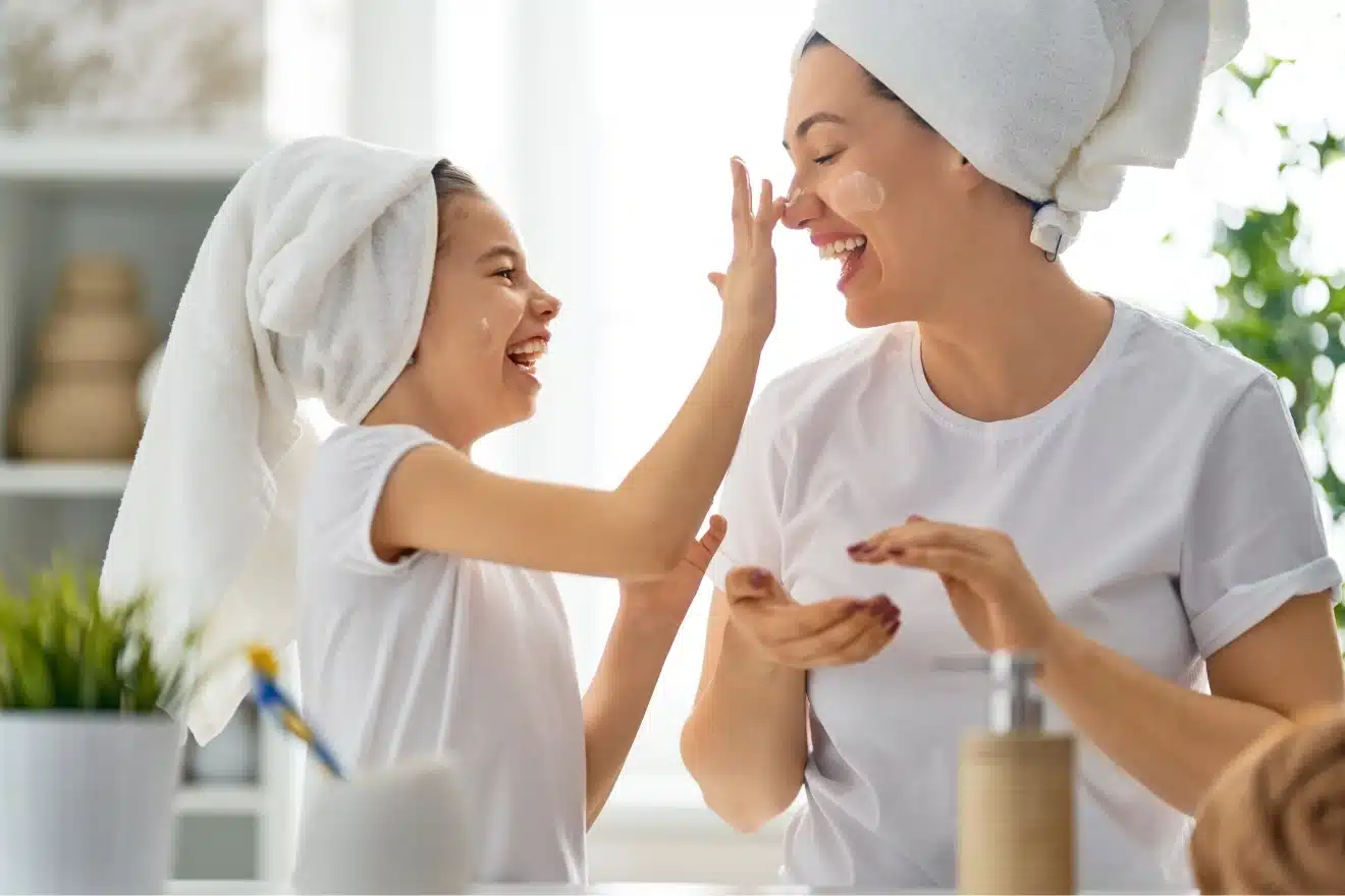A woman and a girl with towels on their heads apply cream to each other’s faces and smile while sitting at a table in a bright room.