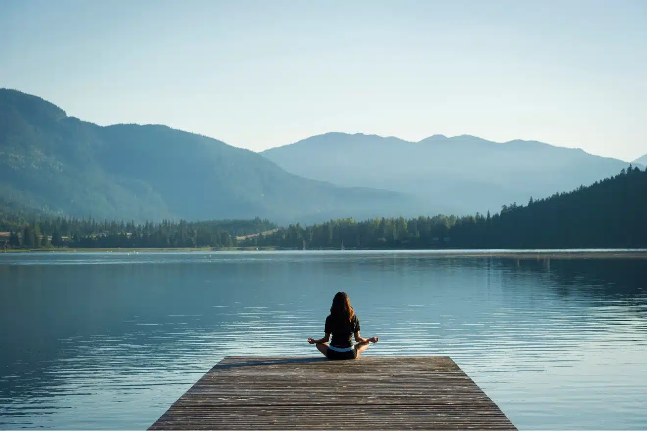 A person sits cross-legged on a wooden dock overlooking a calm lake and distant forested mountains under a clear sky.