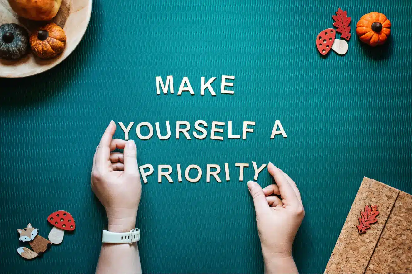 Two hands arrange wooden letters on a teal surface to spell out "MAKE YOURSELF A PRIORITY," with autumn-themed decorations around the edges.