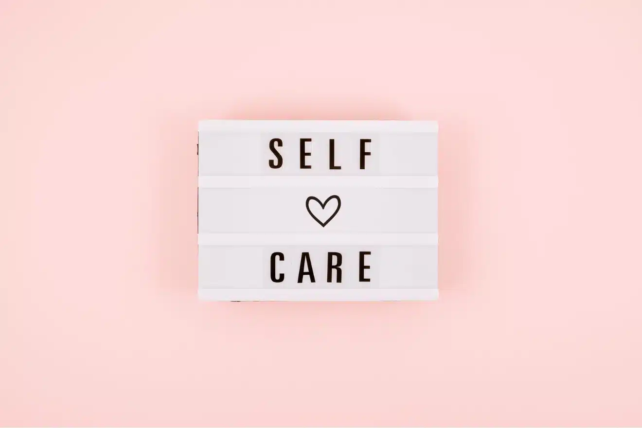 A lightbox sign on a pink background displays the words "SELF CARE" with a heart symbol between the words.