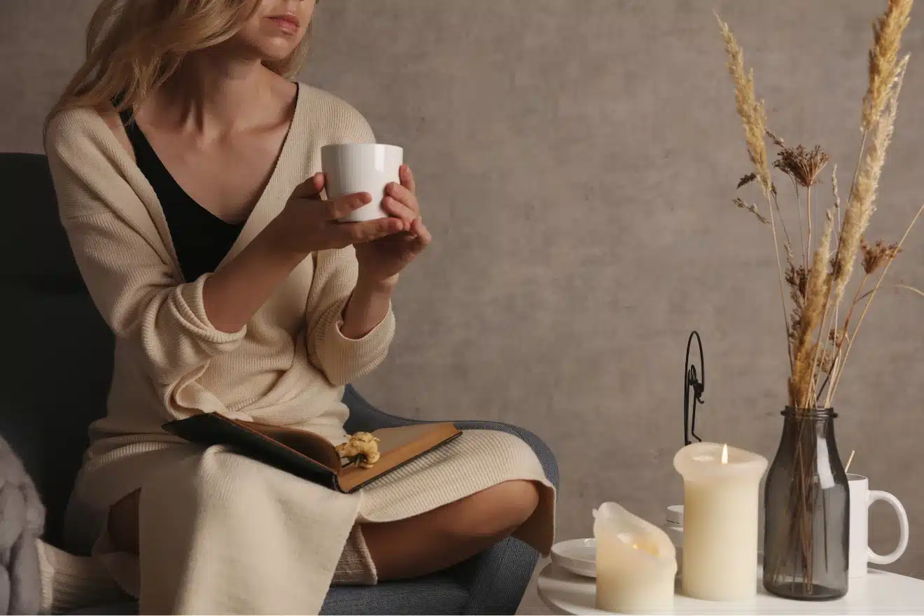 A woman in a cream robe sits with a book and a cup, next to a table holding candles, a teapot, and a vase with dried grasses.