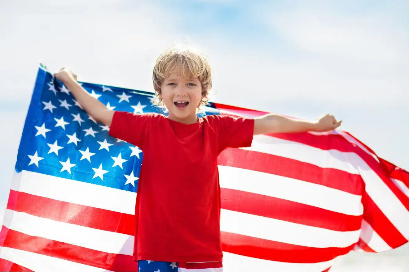 Smiling child in a red shirt stands outdoors holding a large American flag spread out behind them.