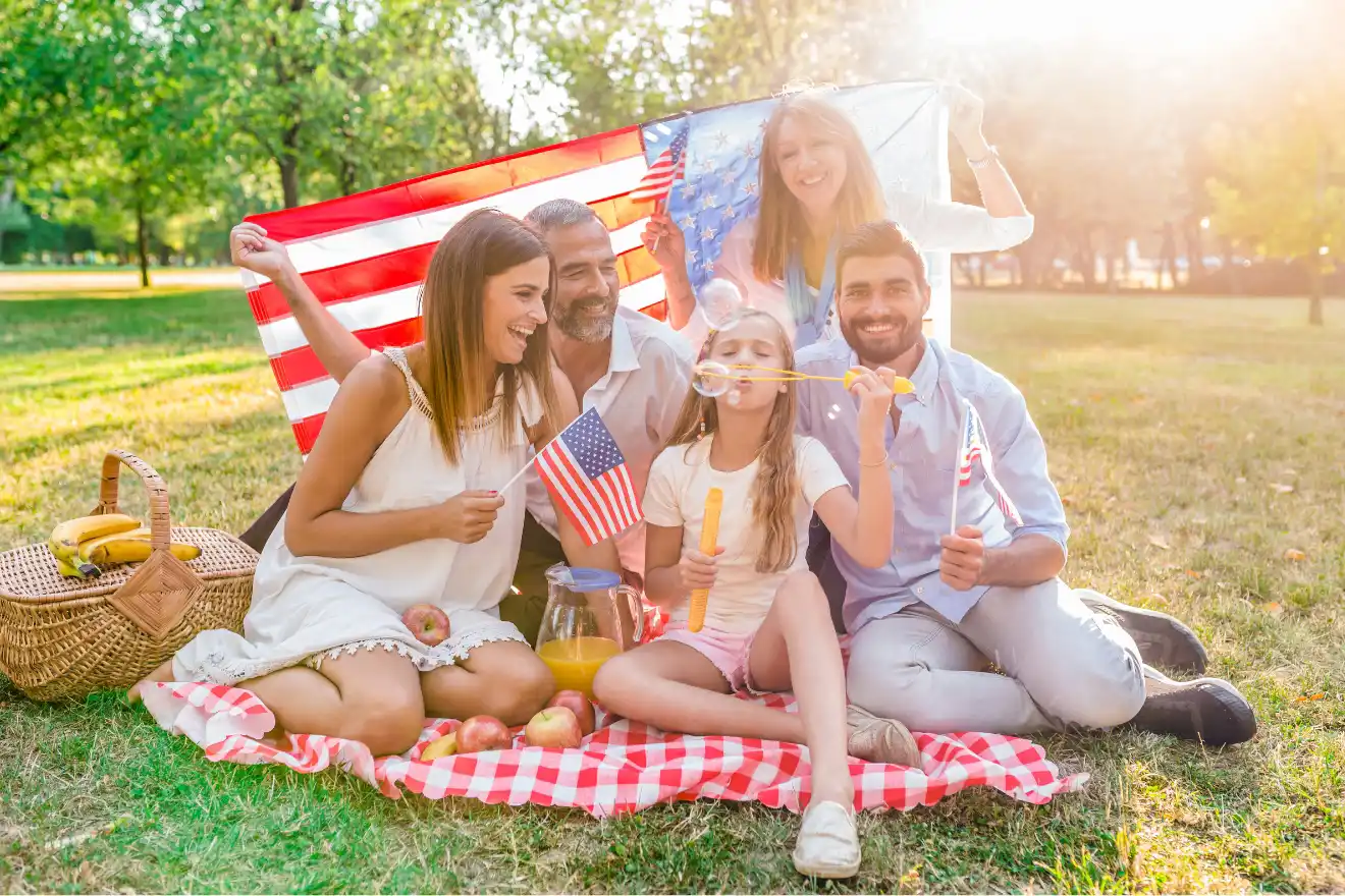 Five people sit on a picnic blanket with an American flag, smiling and holding flags and bubble wands; a picnic basket and food are nearby on the grass in a sunlit park.