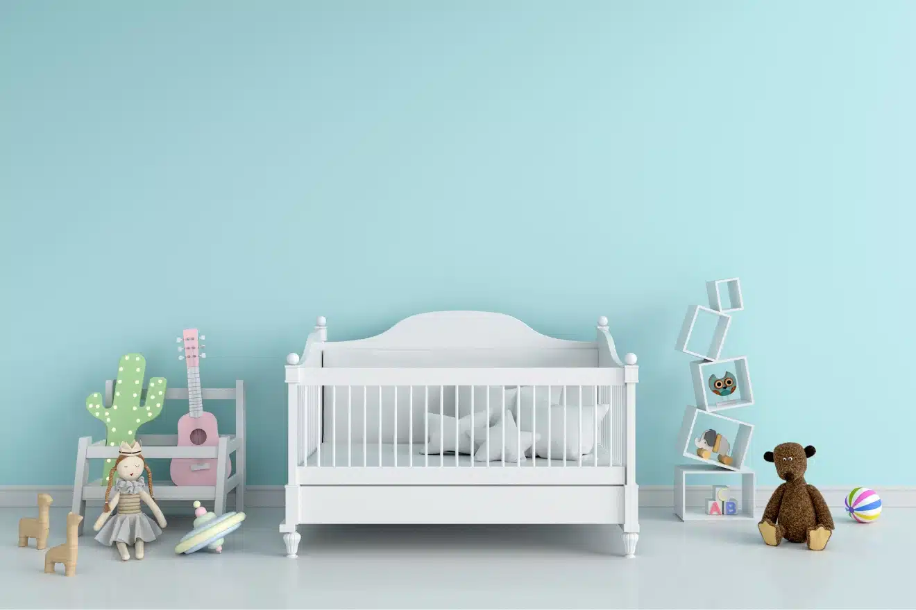 A white baby crib sits against a light blue wall, surrounded by toys, a teddy bear, a pink guitar, and shelves with books and decorations.