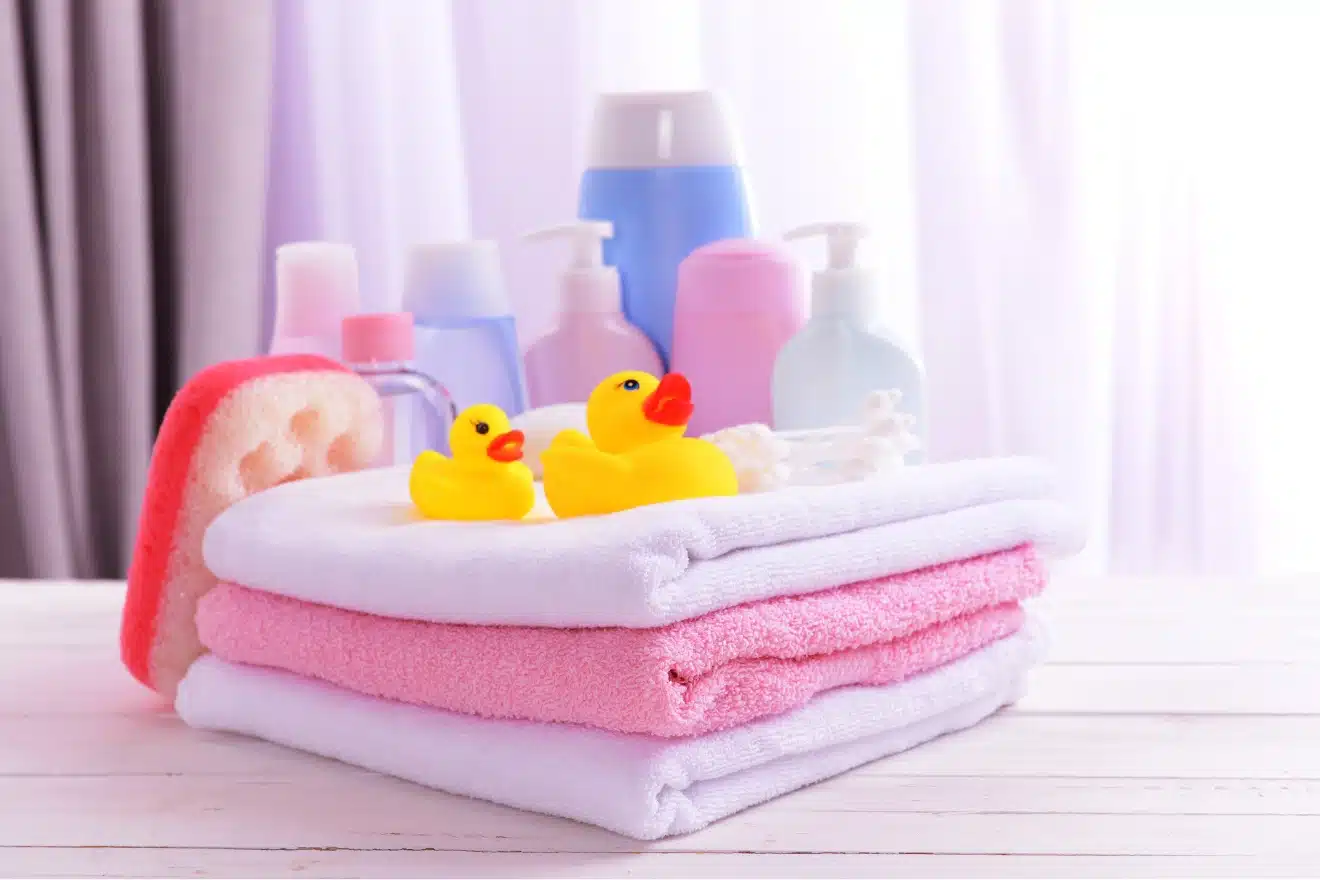 Stacked pink and white towels with two yellow rubber ducks, a sponge, and various bottles of toiletries in the background on a white surface.