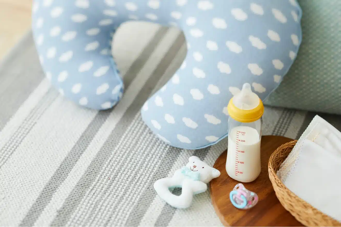 A nursing pillow, baby bottle with milk, soft toy, pacifier, and cloth napkins are arranged on a striped fabric surface.