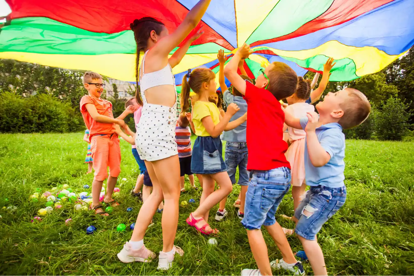 A group of children play outdoors on grass, holding up a large, colorful parachute together.