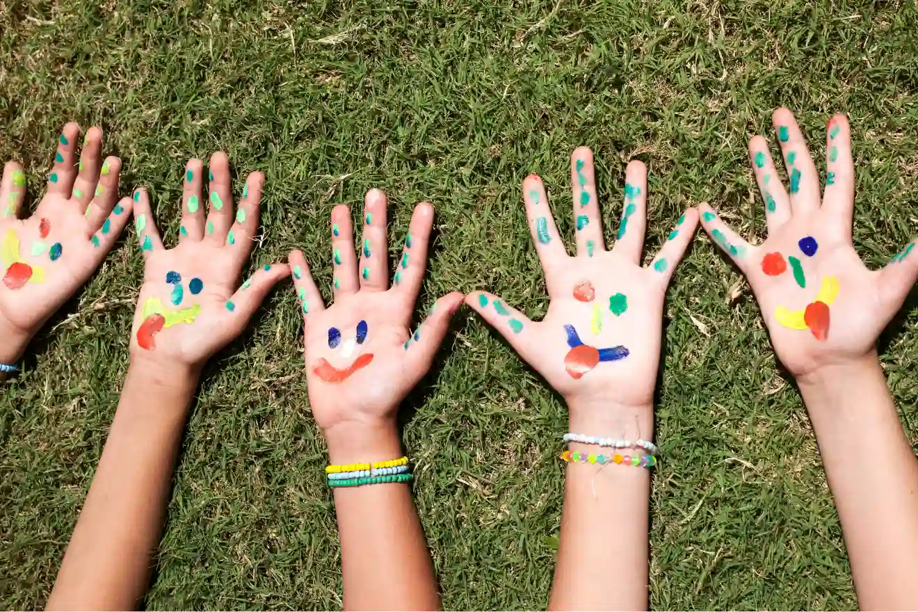 Four hands with colorful painted designs and smiley faces are held open on green grass, with beaded bracelets visible on two wrists.