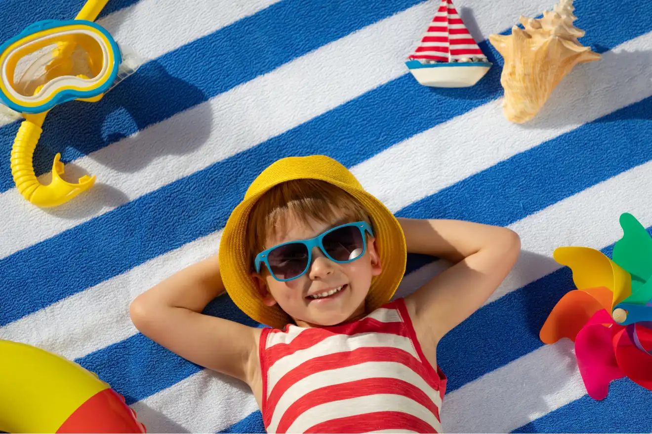 A child in sunglasses and a yellow hat lies on a striped beach towel, surrounded by beach toys, a pinwheel, and a toy sailboat, smiling at the camera.