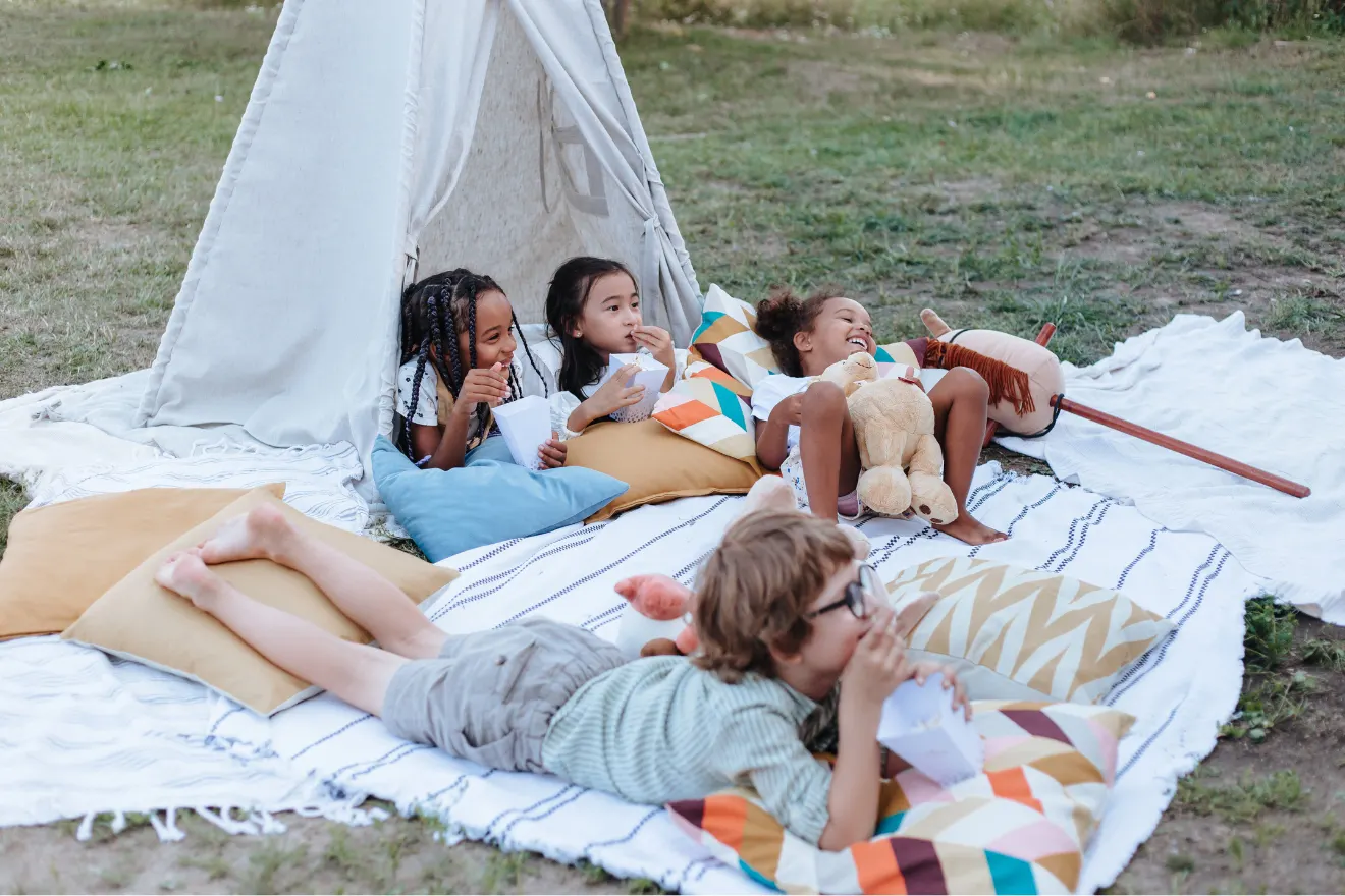 Four children relax on blankets and pillows outside a small tent, eating snacks and lying on the grass with a stuffed toy and wooden stick nearby.