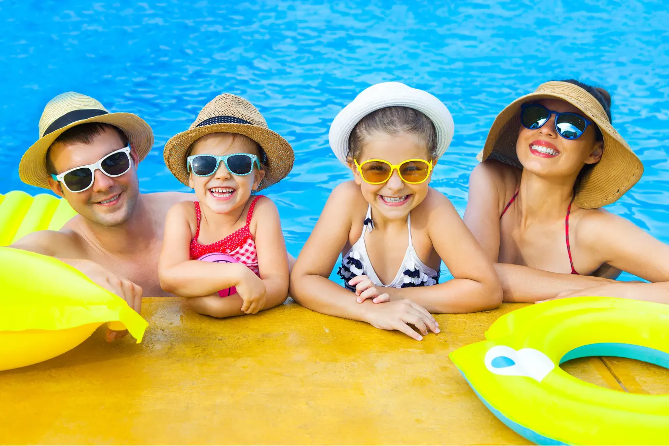 Two adults and two children wearing hats and sunglasses smile while leaning on the edge of a swimming pool, with inflatable pool floats beside them.