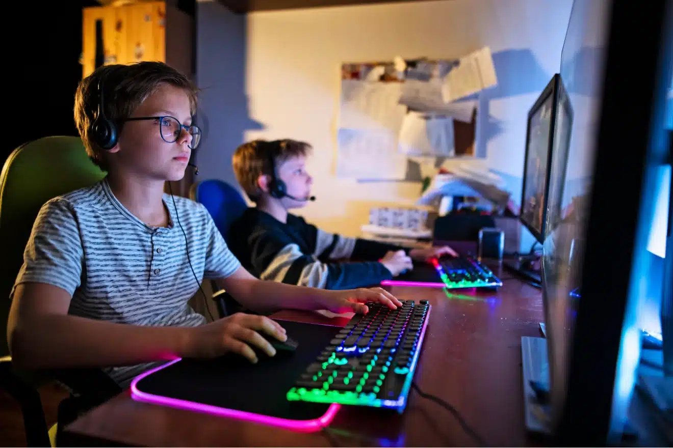 Two boys wearing headsets sit at a desk, using computers with illuminated keyboards and mice, focused on their screens in a dimly lit room.