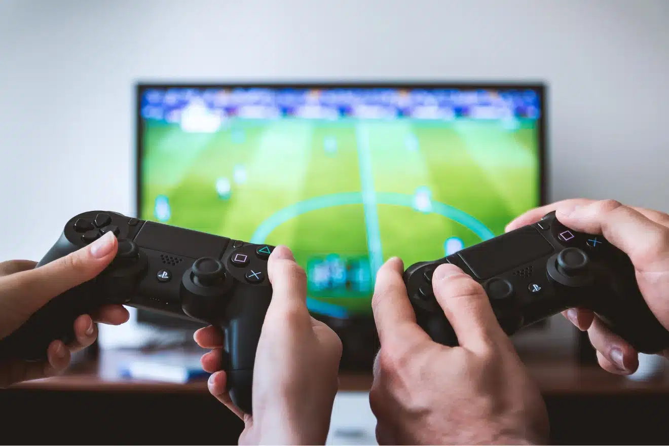 Two people holding game controllers, playing a soccer video game on a television screen in the background.