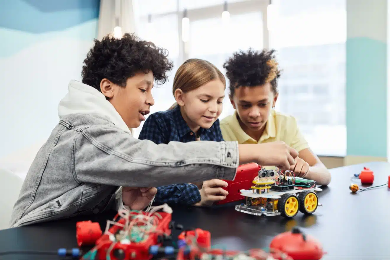 Three children work together on assembling a small wheeled robot at a table, with electronic components and tools scattered nearby.
