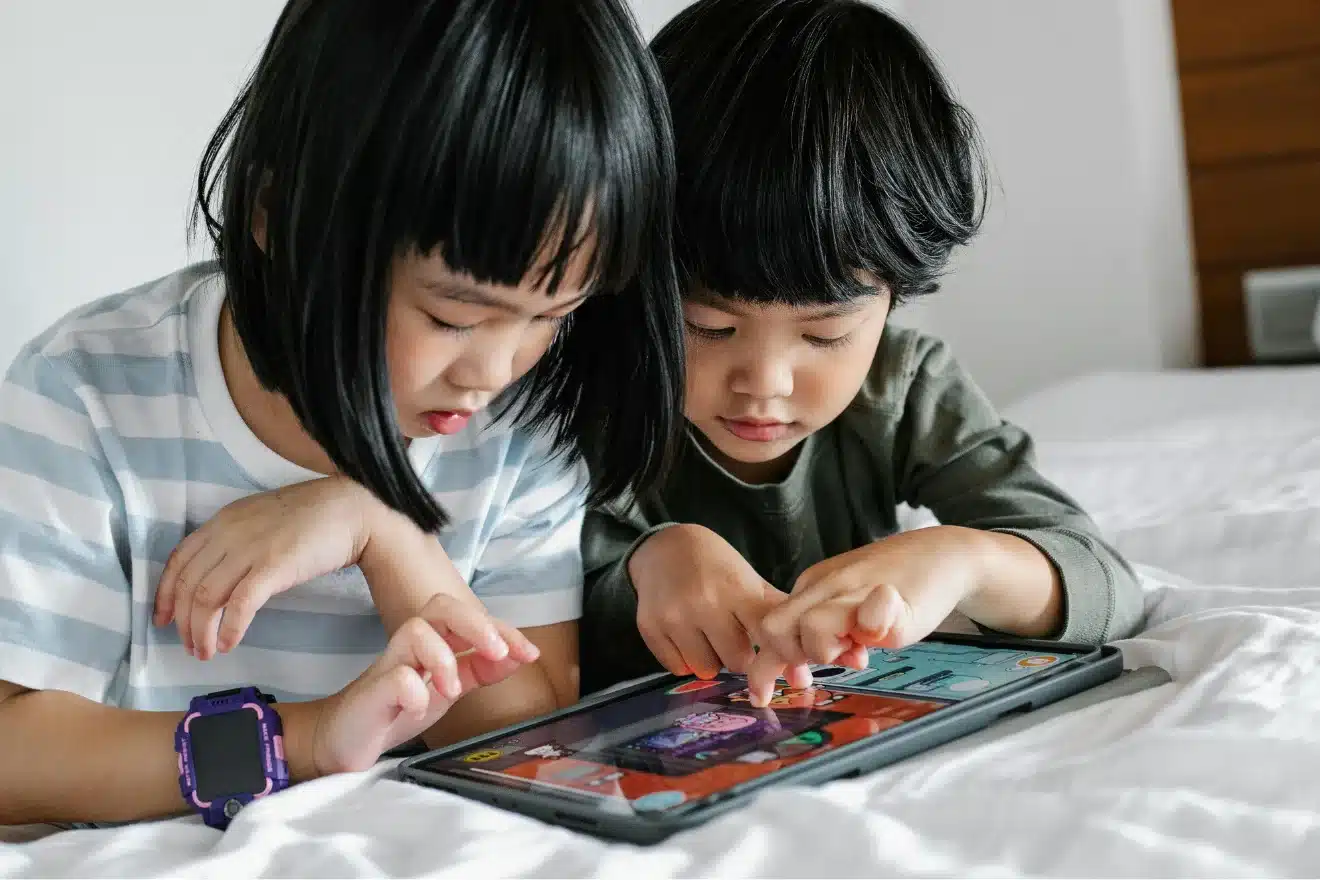 Two young children sit on a bed focusing on a tablet, both using their fingers to interact with a game or application on the screen.