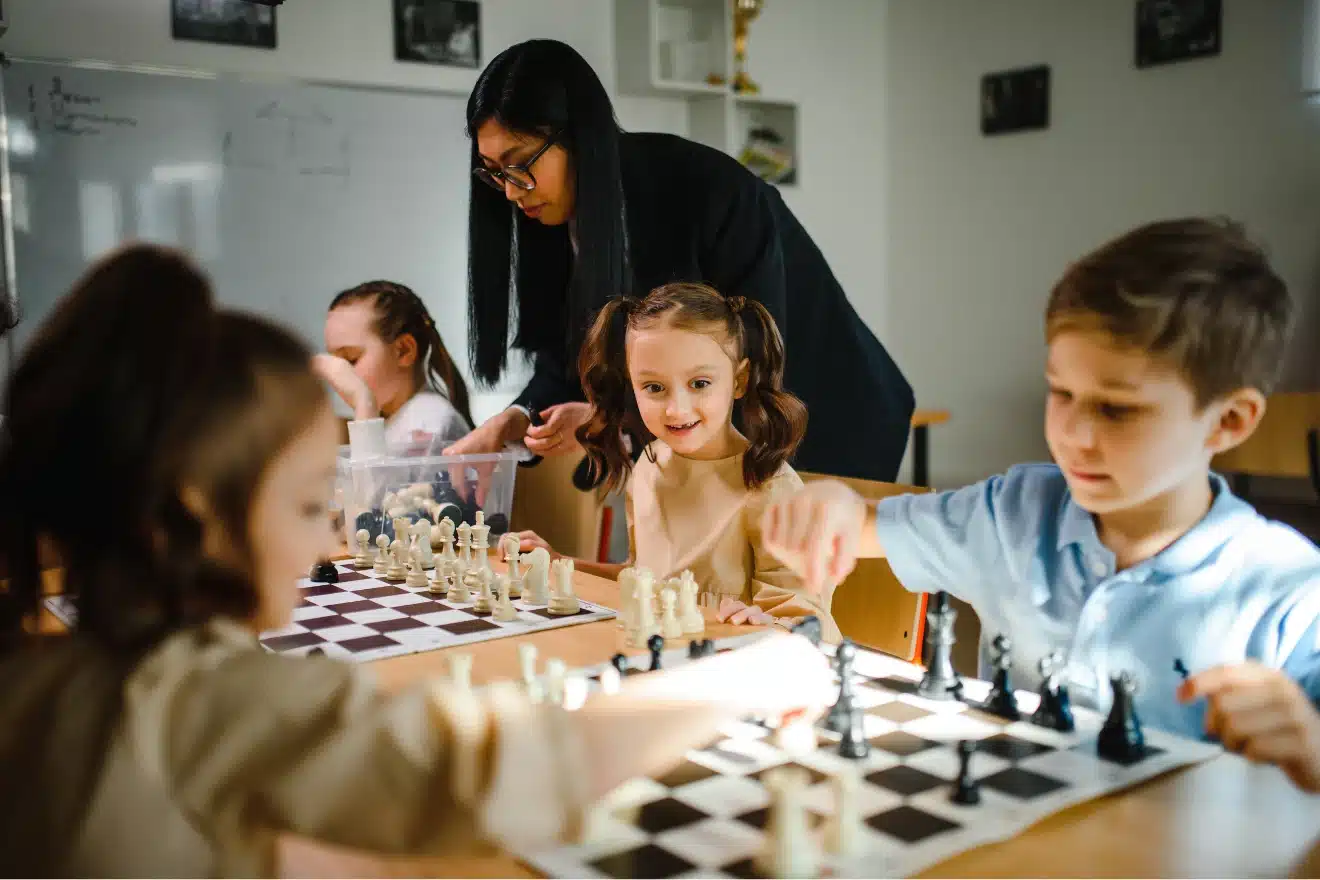 A teacher supervises young children playing chess at a classroom table, as they focus on their chessboards and pieces.