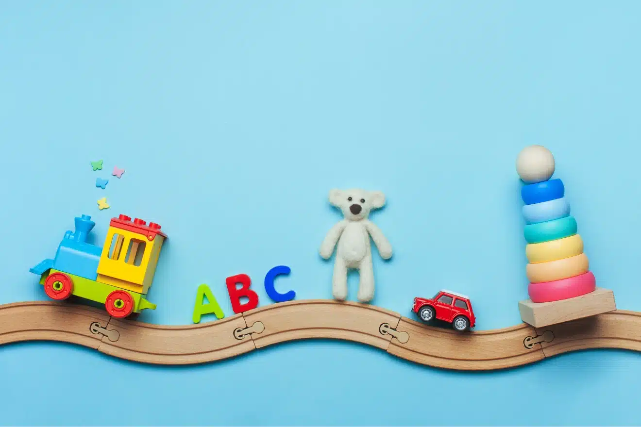 Children's toys including a train, ABC letters, a teddy bear, a red toy car, and a stacking ring tower are arranged on a wooden track against a blue background.