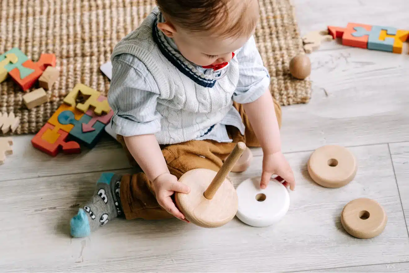 A young child sits on the floor, playing with a wooden stacking toy. Colorful puzzle pieces and blocks are scattered nearby on a rug.