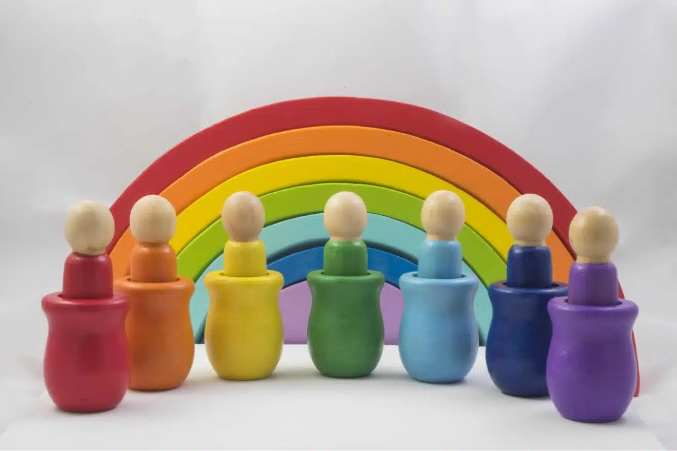 Seven wooden peg dolls in rainbow-colored cups are arranged in front of a wooden rainbow arch, with each doll matching the color of the arch segment behind it.