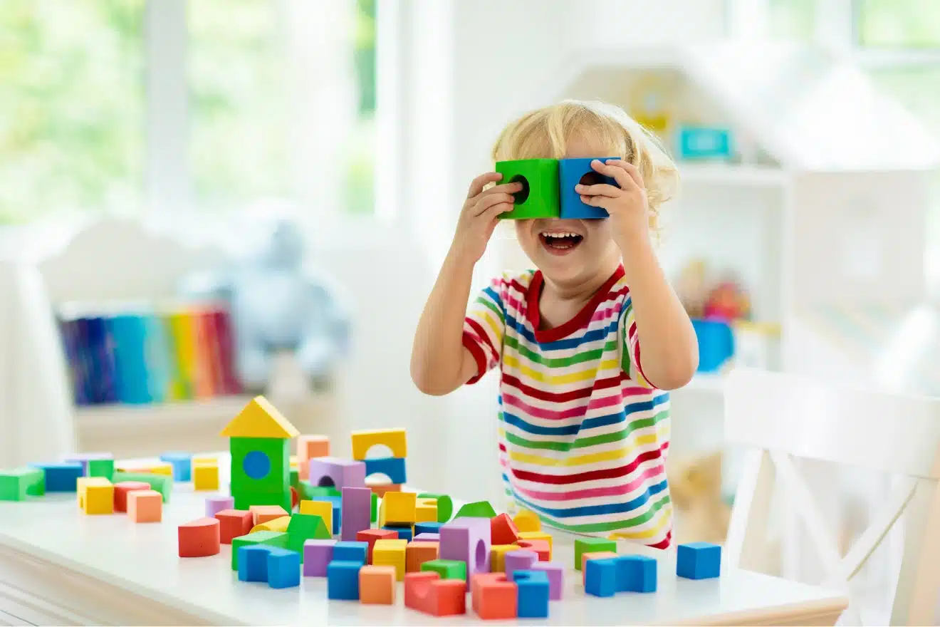 Child in a striped shirt playing at a white table with colorful wooden blocks, holding two blocks up to their eyes and smiling in a bright, tidy room.