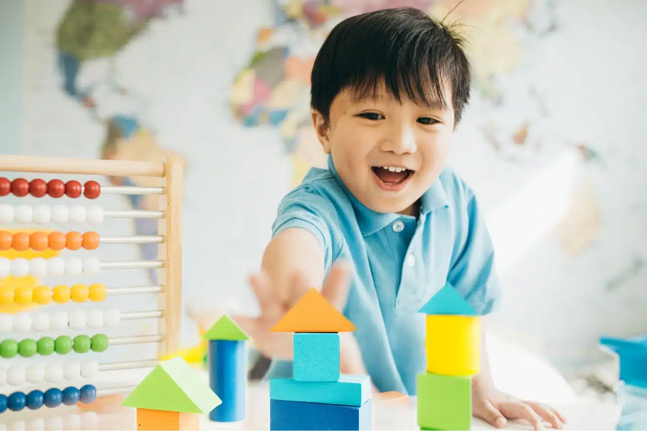A young boy in a blue shirt smiles while playing with colorful wooden blocks and an abacus, with a world map in the background.