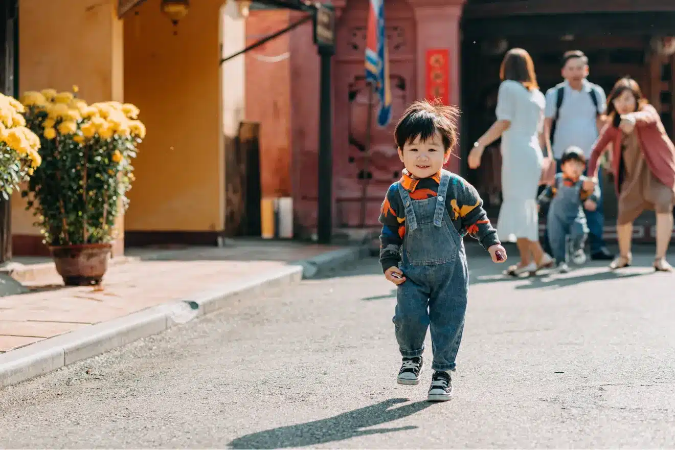 A young child in overalls walks on a sunlit street while several adults and children stand and interact in the background.