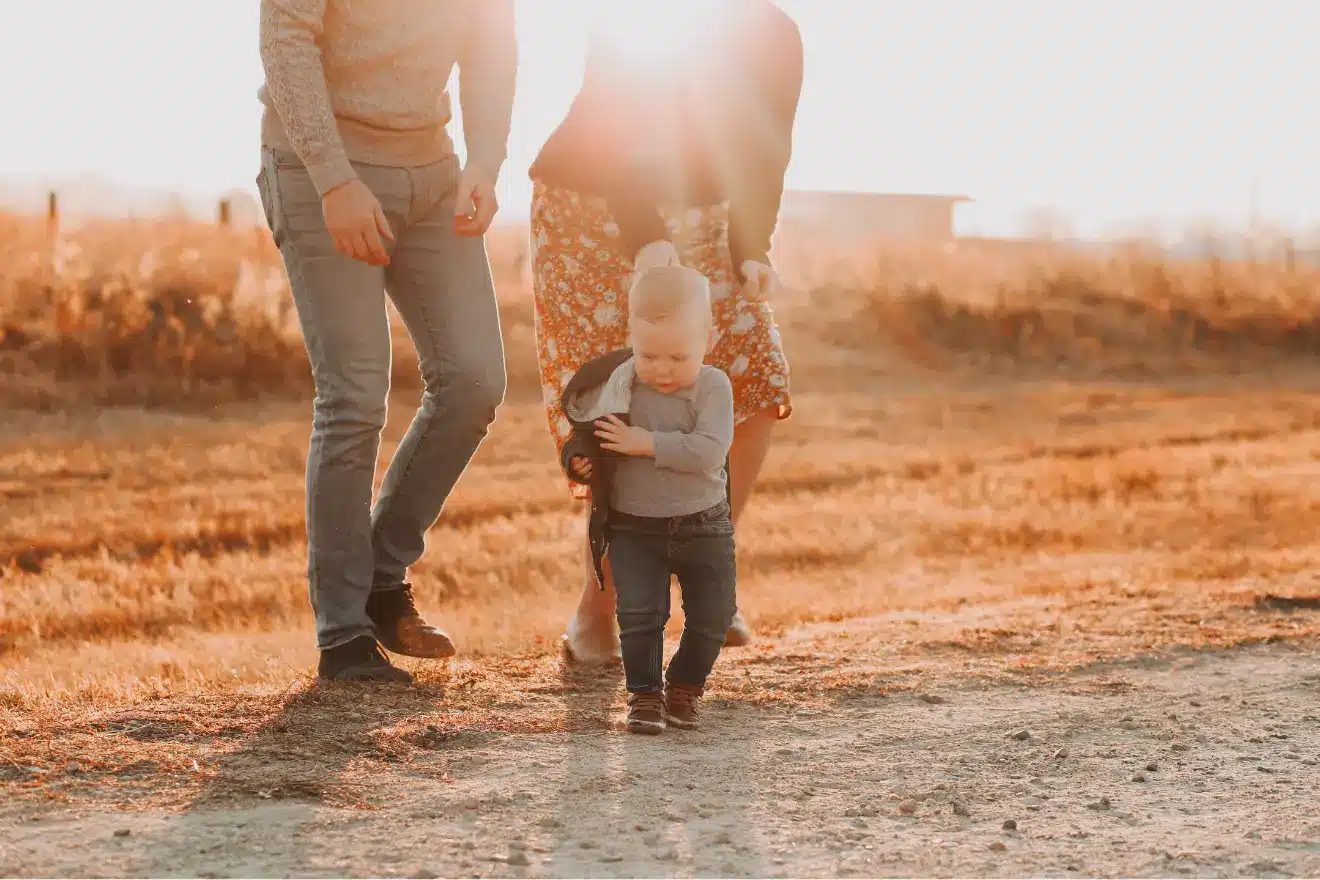 A toddler walks on a dirt path outdoors, holding a small jacket, with two adults nearby in a sunlit, rural setting.
