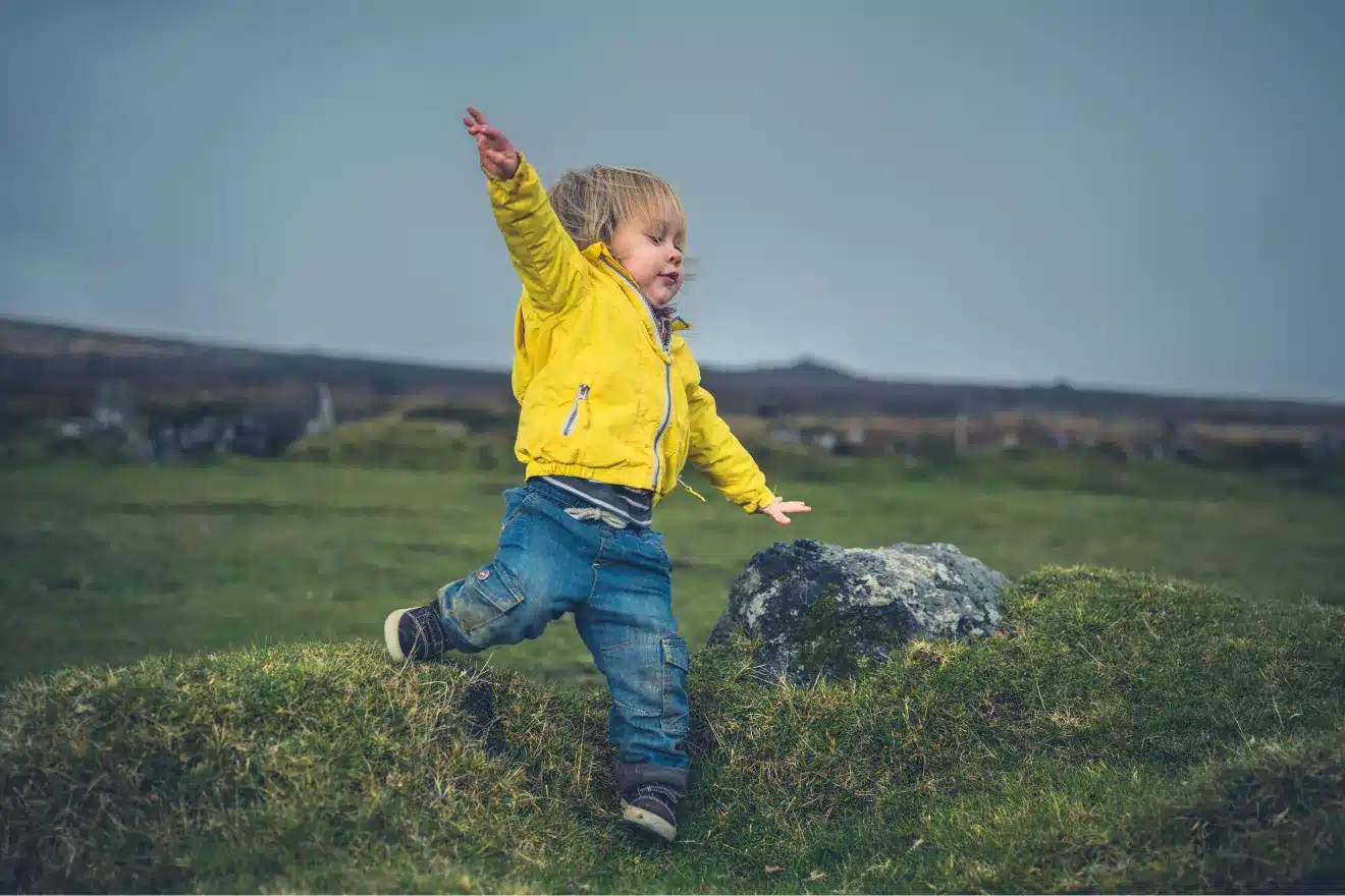 A young child in a yellow jacket and jeans balances with arms outstretched while walking on grassy terrain outdoors, with rocks and an overcast sky in the background.