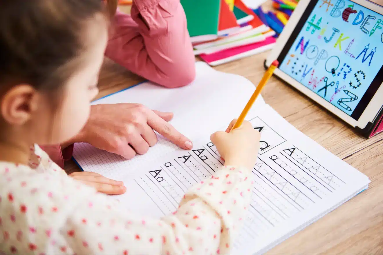 Child practicing writing the letter "A" in a workbook with guidance from an adult, while an alphabet chart is displayed on a tablet nearby.