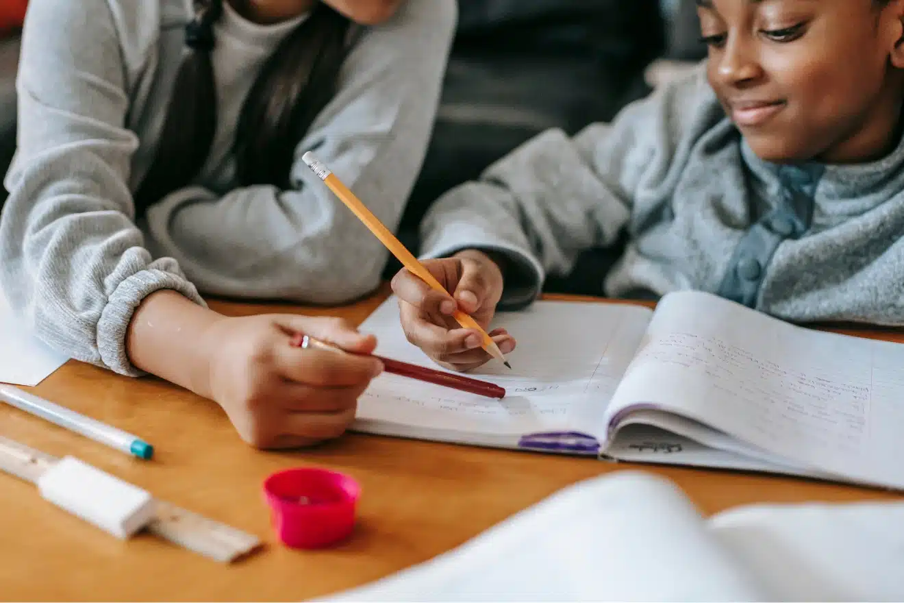 Two children sit at a table working together on a notebook, with pencils, a ruler, and other school supplies scattered nearby.