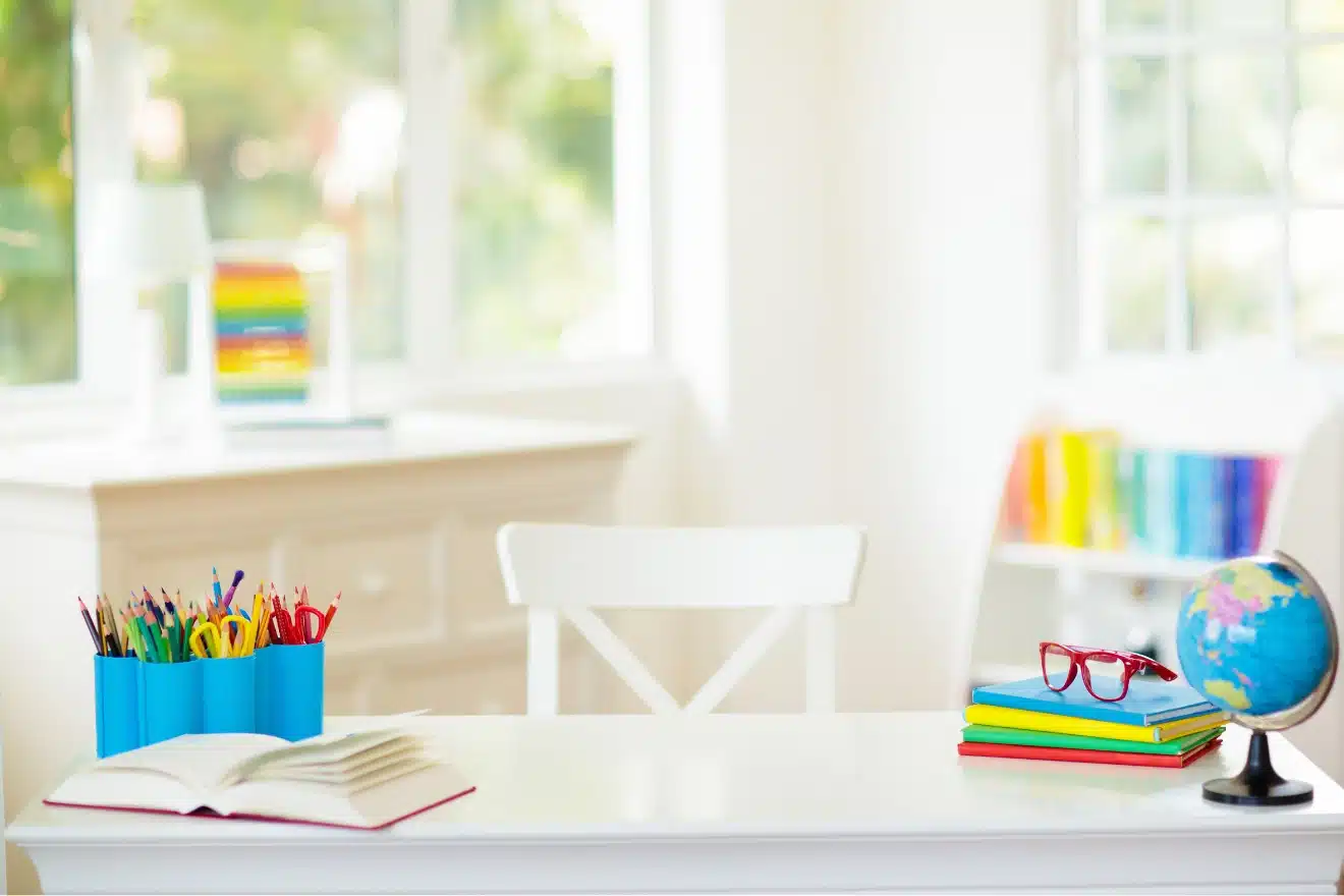 A white desk with colored pencils, an open book, stacked notebooks with red glasses, and a small globe, set in a bright room with bookshelves.