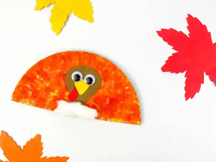 A paper plate turkey craft with googly eyes, a red and yellow beak, and cotton at the bottom, surrounded by cut-out paper leaves on a white background—perfect for Thanksgiving crafts with kids.