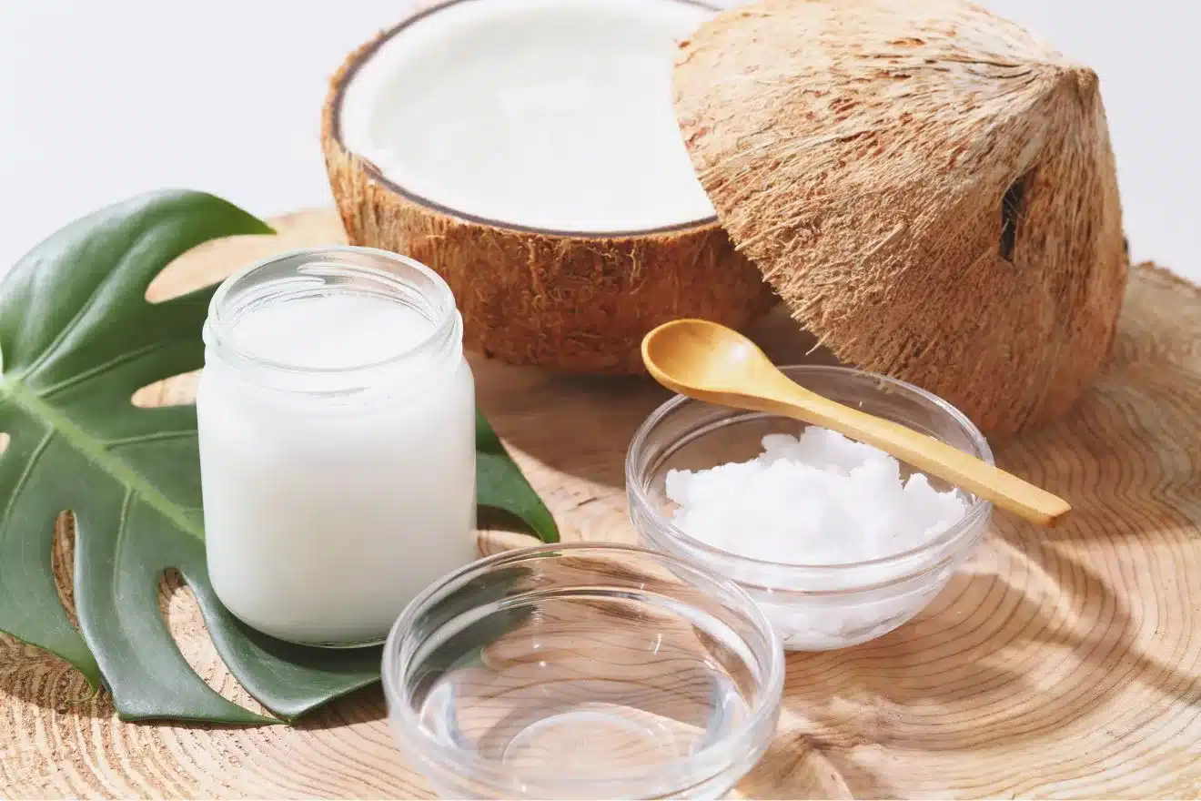 A halved coconut, a jar of coconut oil, a leaf, two glass bowls—one empty, one with solid coconut oil—and a small wooden spoon arranged on a wooden surface.