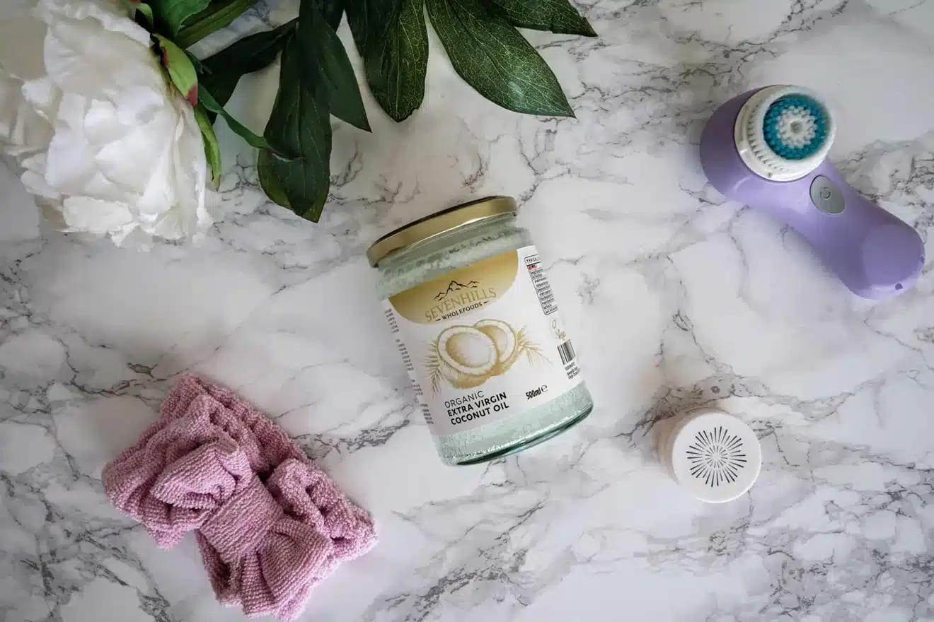 A jar of organic virgin coconut oil lies on a marble surface near a purple facial brush, a pink cloth, a white flower, and green leaves.