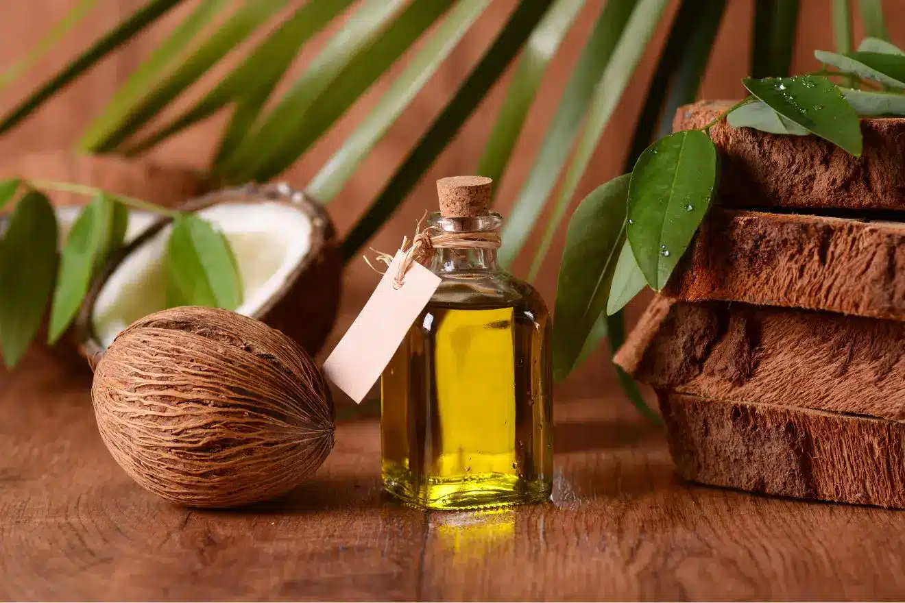 A small glass bottle of yellow oil with a cork and tag sits on a wooden surface next to a whole coconut, a split coconut, stacked wood, and green leaves.