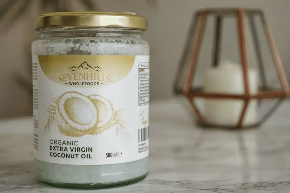 A jar of Sevenhills Wholefoods organic extra virgin coconut oil sits on a marble surface with a decorative candle holder in the background.