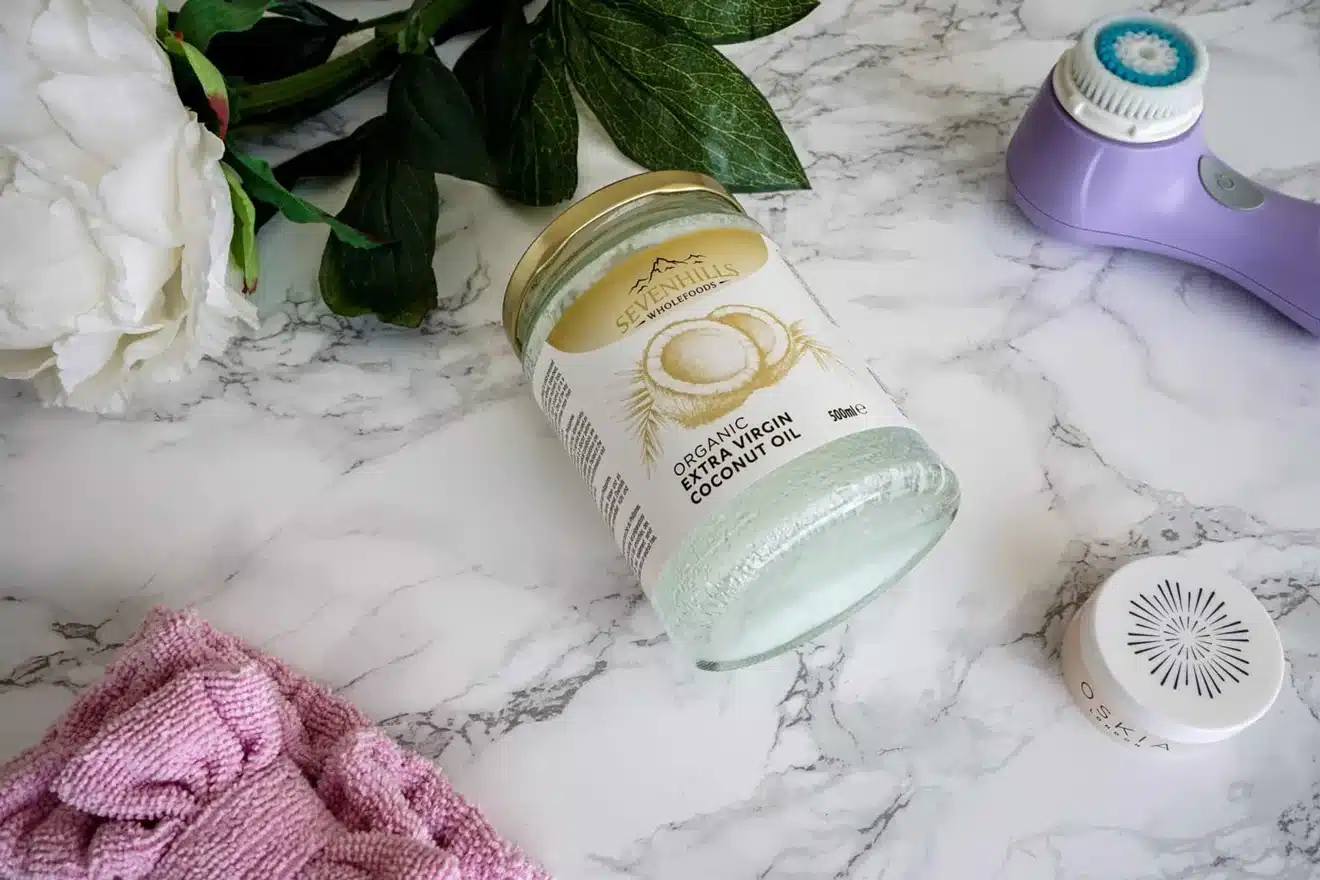 A jar of organic virgin coconut oil lies on a marble surface near a purple facial brush, a pink cloth, a white flower, and green leaves.