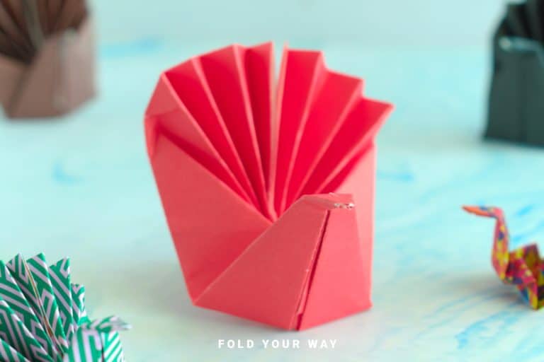 A red origami turkey made from folded paper is displayed upright on a light blue surface, with other origami figures in the background—perfect for Thanksgiving crafts or fun holiday crafts with kids.