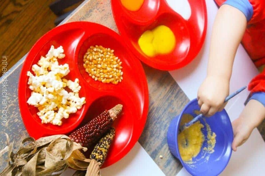 A child mixes batter in a blue bowl near two red trays filled with eggs, popcorn kernels, popped popcorn, and colorful corn on the cob—perfect inspiration for Thanksgiving crafts for kids.