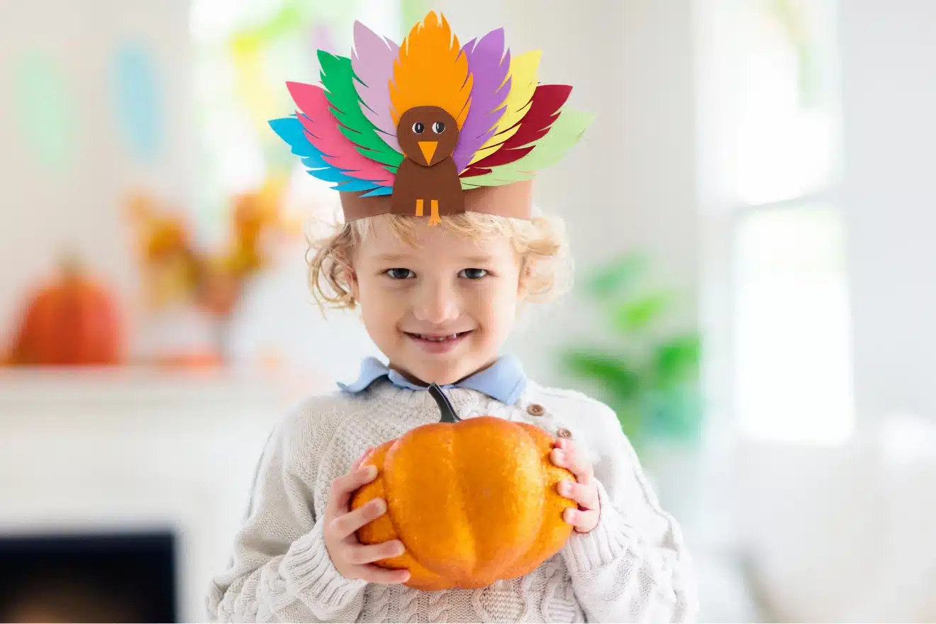 A young child wearing a colorful turkey feather headband smiles while holding a small pumpkin indoors.