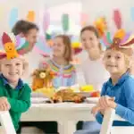 Two young children wearing colorful paper turkey hats sit at a table set with food, while two adults and another child—perhaps gearing up for Thanksgiving Day family games—are blurred in the background.