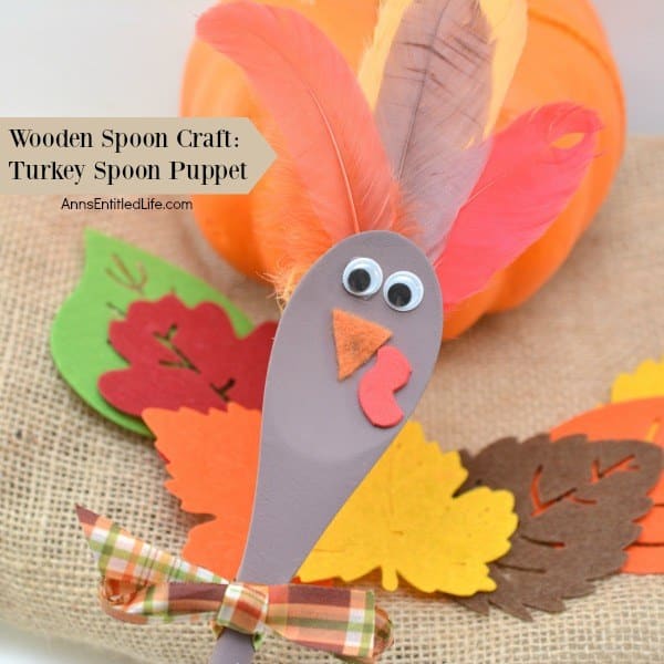 A wooden spoon decorated as a turkey puppet with googly eyes, felt beak, paper wattle, feathers, and a plaid bow—surrounded by felt leaves and a pumpkin—makes a festive Thanksgiving crafts project and fun holiday idea for kids.
