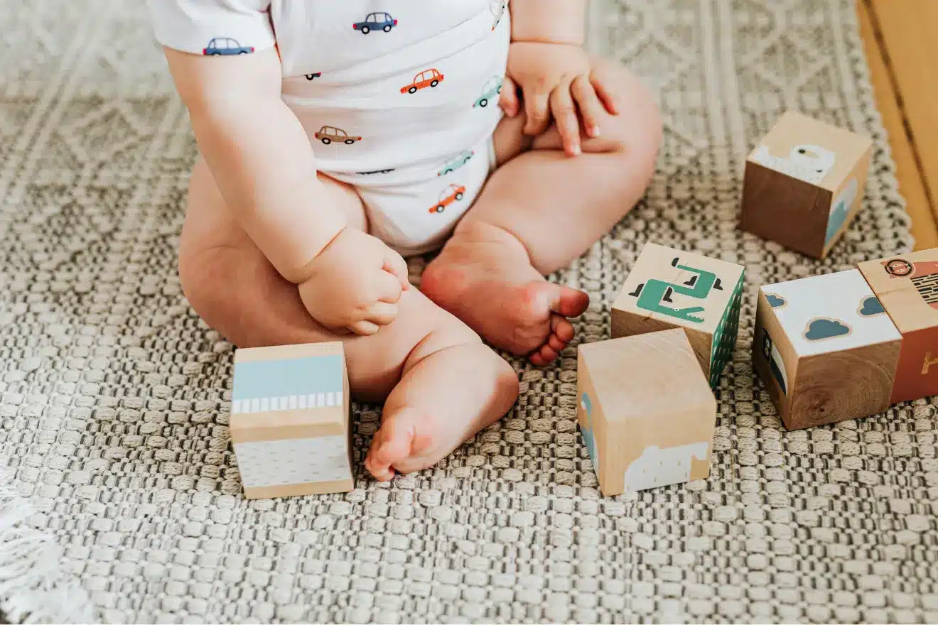 A baby in a white onesie sits on a patterned rug, playing with colorful wooden blocks decorated with various designs.